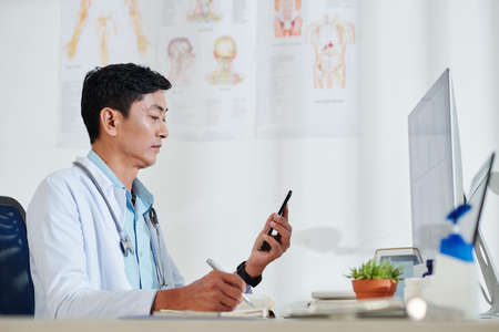 Serious mature Vietnamese doctor checking text messages from patients and writing down his complaintsの写真素材