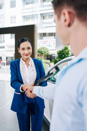 Pretty smiling young sales manager shaking hand of customer, greeting him before showing carsの写真素材