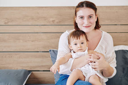 Portrait of cheerful young mother sitting with baby girl on her laps and looking at cameraの写真素材