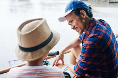 Happy mature Asian man spending weekend with son on lake, fishing and talkingの写真素材