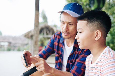 Father showing photo of fish on smartphone screen to preteen son when they are fishing in lake...