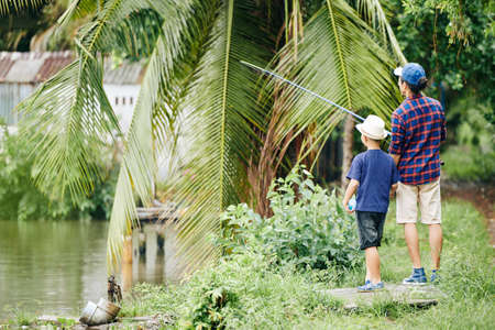 Little boy in hat looking at his father fishing in lake with new rod, view from the backの写真素材