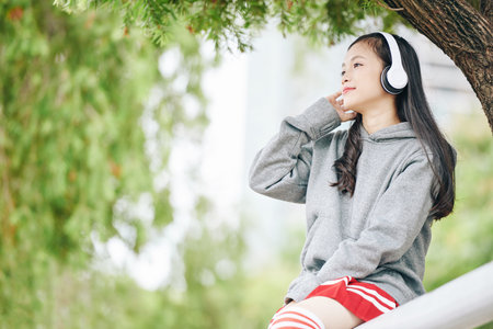 Smiling charming young Vietnamese girl spending time in park and listening to music in headphone