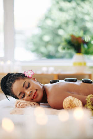 Charming young African-American woman lying on bed in spa salon with hot basalt stones on her backの写真素材