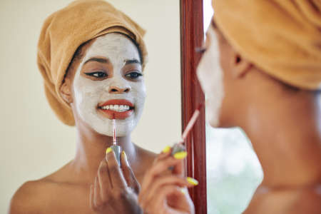 Smiling attractive young African-American woman with towel on her head applying liquid lipstick when getting ready in the morningの写真素材