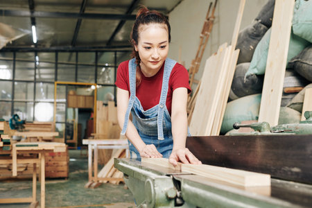 Smiling young Asian female carpenter cutting wooden board into planks