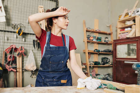Young female carpenter wiping sweat after working with wood in carpentry shopの写真素材