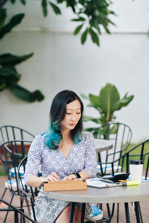 Pretty young Chinese businesswoman sitting at cafe table, drinking coffee and working with reports...