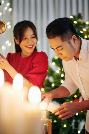 Pretty smiling young Asian woman looking at her boyfriend burning candles when decorating apartment for Christmasの写真素材