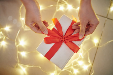 Hands of young woman opening Christmas present, glowing lights in background