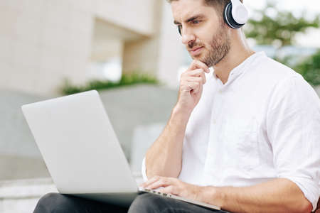 Pensive young freelance software developer rubbing chin and looking at laptop screenの写真素材