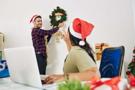 Businesswoman in Santa hat asking colleague to hang Christmas wreath on office wallの写真素材