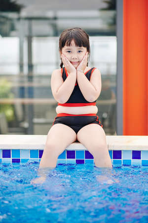 Cute Asian little girl sitting on pool edge, touching her cheeks and looking at cameraの写真素材