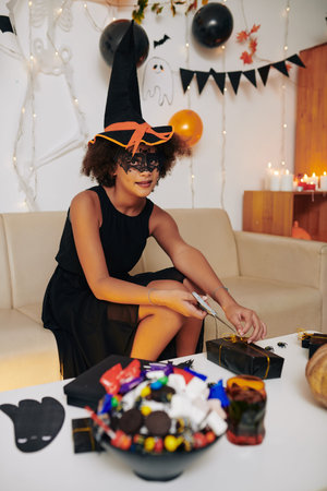 Girl in Halloween witch costume wrapping presents and decorating room for celebration