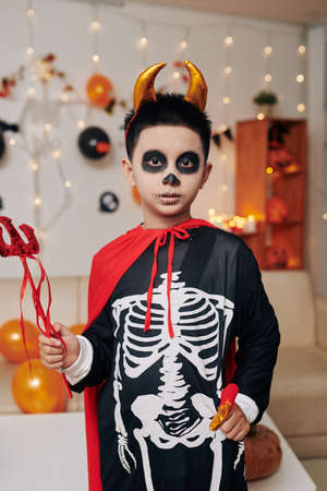 Confident little boy dressed as devil holding trident and looking at camera for Halloween photoの写真素材