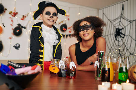 Portrait of happy excited brother and sister in Halloween costumes standing at table with party snacks and drinksの写真素材