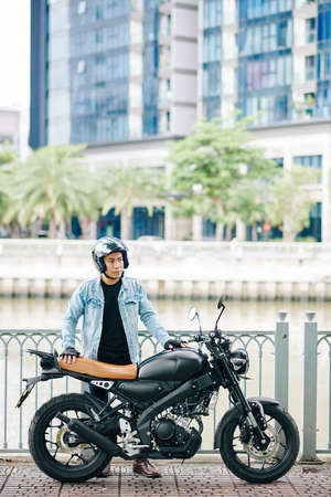 Young serious Asian man in helmet standing on bridge next to his motorcycleの写真素材