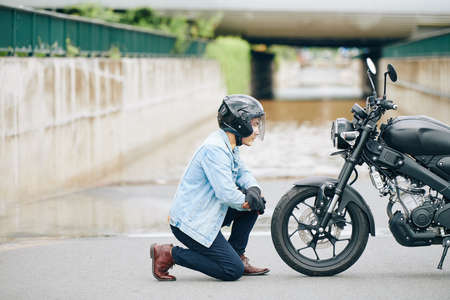 Serious Asian motorcyclist standing on one knee and looking at headlightの写真素材