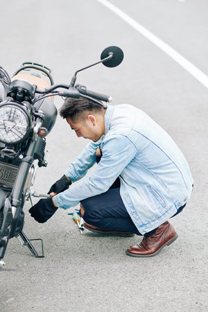 Serious Asian man inspecting frame, suspension and chain of motorcycle before riding