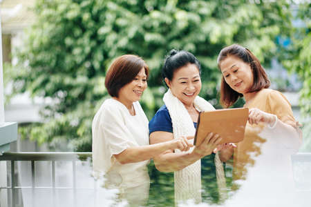 Cheerful senior Vietnamese female friends using new application on tablet computer, discussing photos or newsの写真素材