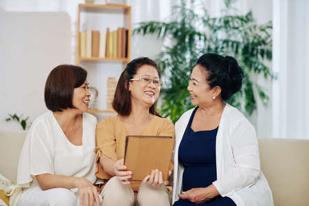 Group of happy Vietnamese elderly women watching funny videos or looking at photos on tablet computer when meeting at homeの写真素材