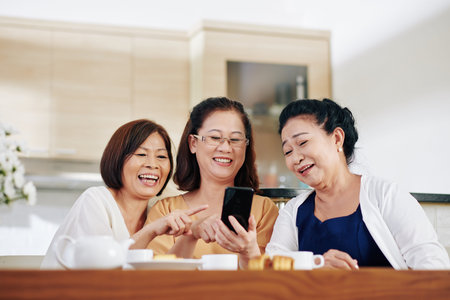 Group of cheerful Vietnamese senior women watching funny videos on smartphone screen