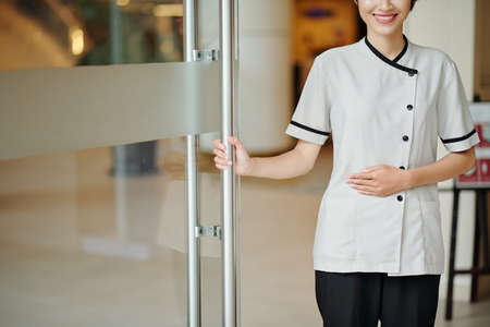 Cropped image of smiling young hotel manager in uniform opening glass entrance doorsの写真素材