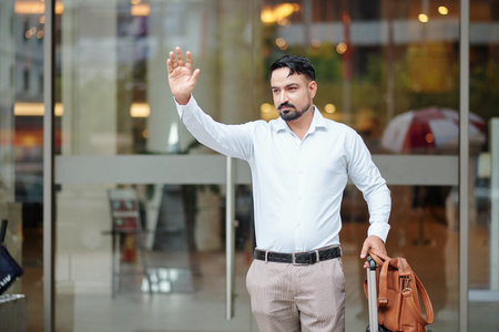 Handsome hotel guest with bag and suitcase leaving hotel and catchig taxi