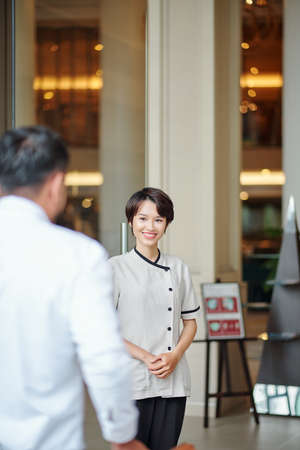 Smiling young Asian concierge looking at man entering hotel lobbyの写真素材