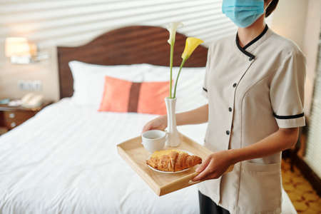 Hotel maid wearing medical mask when bringing tray with breakfast and flowers in hotel roomの写真素材