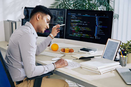 Pensive Indian developer checking structure chart when working on programming code at his deslの写真素材