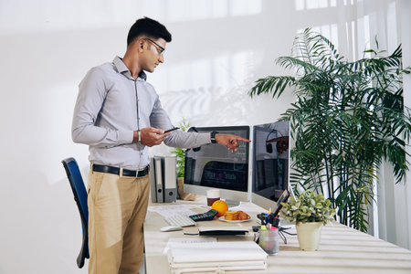 Pensive Indian programmer with smartphone in hand pointing at computer screen with programming code...