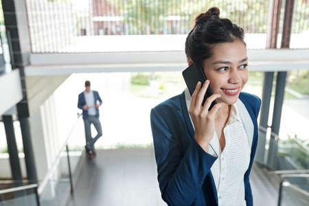 Positive young pretty business lady standing in office building and talking on phoneの写真素材