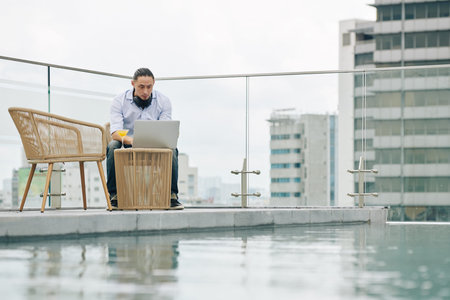 Serious young man sitting by swimming pool on rooftop with glass of juice and working on laptop