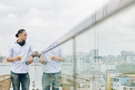 Pensive young mixed-race man standing on rooftop with glass of juice and smartphone and enjoying...
