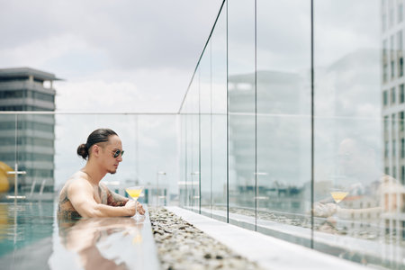 Serious young man in sunglasses spending time in swimming pool on rooftop and enjoying city view