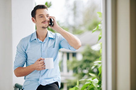 Portrait of positive handsome young man drinking morning coffee and talking on phone with friend