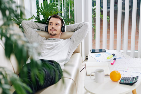 Young man relaxing on sofa and listening to music in headphones after working with documents at home