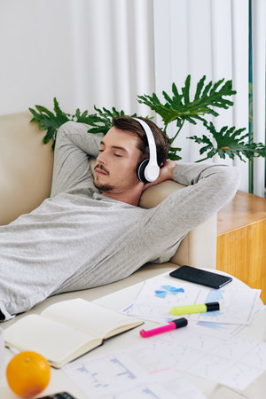 Young businessman in headphones sleeping on sofa at home after checking charts and reports