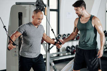 Fitness trainer looking at client doing exercise in cable crossover machine for his upper chest and armsの写真素材