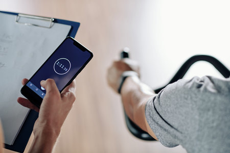 Fitness trainer checking stopwatch on smartphone when his client riding on stationary bike