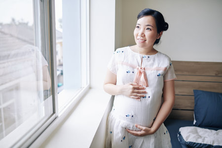 Portrait of cheerful pregnant young Asian woman standing at window in bedroom and touching belly