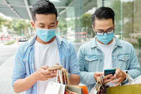 Young Asian men in protective masks standing in the street with shopping bags and texting friends