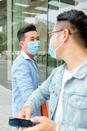 Young Vietnamese men in medical masks greeting each other with elbow bump due to coronavirus...