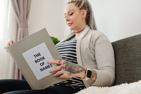 Happy pregnant young woman sitting on sofa at home and reading book of names