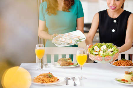 Cropped image of women putting bowl of vegetable salad and plate with spring rolls on dinner tableの写真素材