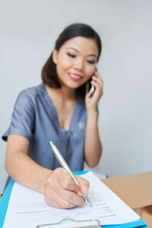 Smiling pretty spa salon or clinic reception worker talking on phone with client and filling questionnaire with his personal data of clientの写真素材