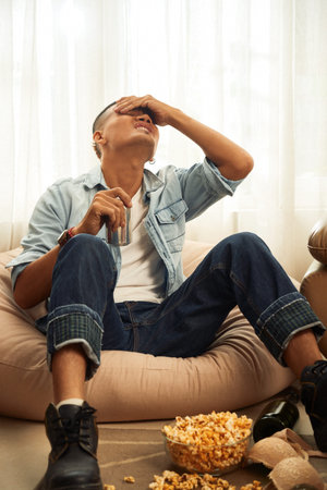Young man suffering from severe headache on the morning after party and drinking can of refreshing...