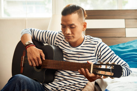 Serious young Vietnamese man spending time in dorm, playing guitar and singing