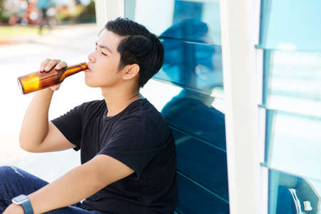 Asian young man drinking beer from bottle while sitting outdoorsの写真素材